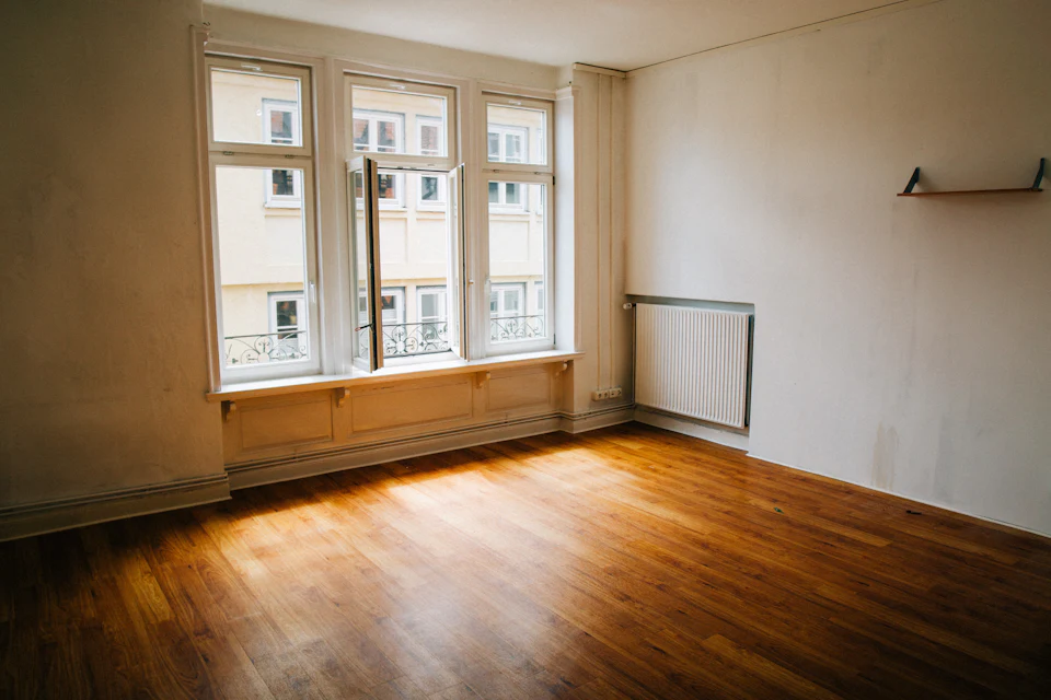 Before staging — Vacant room with dated finishes, warm wood floors and tall windows